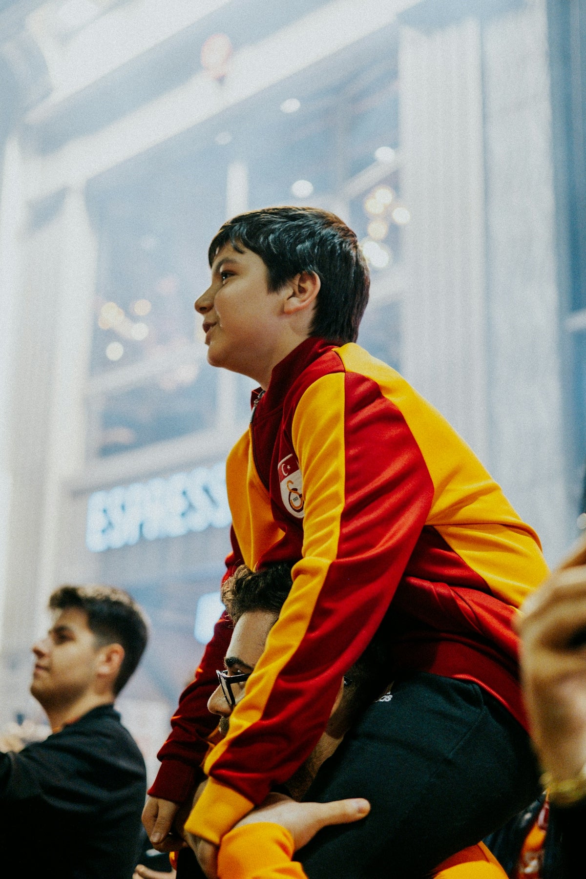 Boy on man's shoulders wearing red and yellow sports jacket.